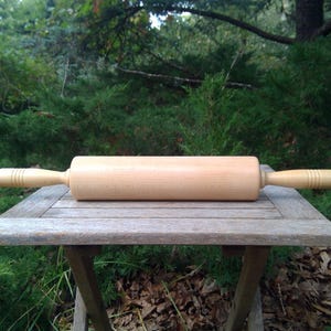 May include: A light-colored wooden rolling pin with a smooth cylindrical barrel and two tapered handles on either side. The handles feature three decorative horizontal grooves carved into the wood near the ends of the central rolling section.