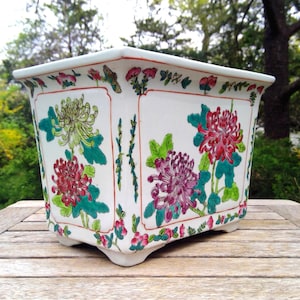 May include: A rectangular white ceramic planter with a floral design. The planter features red, purple, and yellow chrysanthemum flowers with green leaves. The edges are trimmed with red and green floral patterns. The planter sits on four small feet.