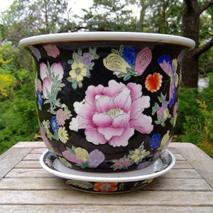 May include: A black ceramic planter with a matching saucer, featuring a vibrant hand-painted floral pattern with large pink peonies, smaller yellow and blue blossoms, and colorful butterflies against a dark background with a white rim.