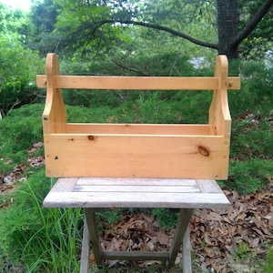May include: A light-colored wooden tote with a handle and a rectangular base. The tote is sitting on a small wooden table outdoors, surrounded by greenery. The tote is made of unfinished wood.