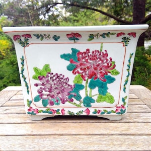 May include: A rectangular white ceramic planter featuring hand-painted floral motifs in shades of red, purple, and green. The design includes large chrysanthemum blossoms and green leaves, framed by a delicate border of small pink flowers and vines.