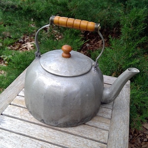 May include: A vintage silver-colored kettle with a wooden handle and lid knob. The kettle sits on a wooden slatted surface, with a green leafy background. The spout is curved and the handle is arched.