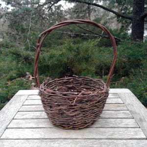 May include: A round, handwoven basket with a handle. The basket is made of brown twigs and sits on a weathered wooden surface. The background features lush green foliage and trees, creating a natural setting.