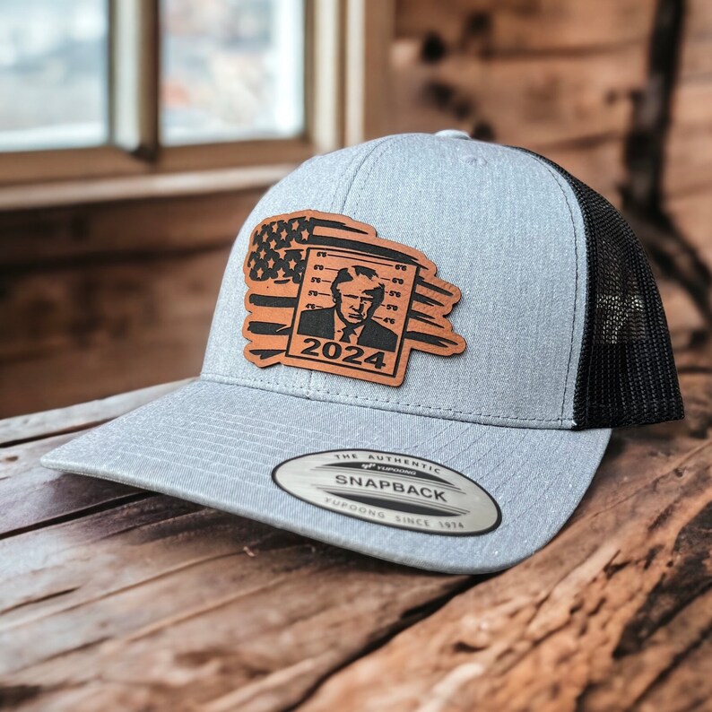 May include: A gray and black baseball cap with a leather patch featuring a portrait of a man with the text "2024" and an American flag design. The patch is attached to the front of the cap. The cap has a black mesh back and a gray front.