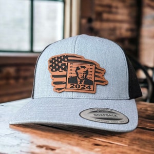 May include: A gray and black baseball cap with a brown leather patch featuring a mugshot of a man with the text "2024" underneath. The patch also includes an American flag design.