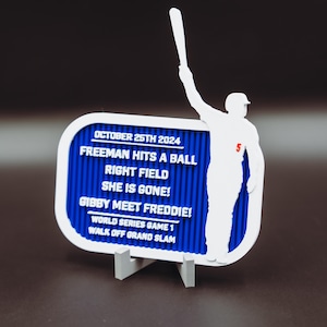Puede incluir: Un letrero blanco y azul con temática de béisbol con la silueta de un jugador de béisbol sosteniendo un bate. El letrero tiene el texto "Freeman batea una bola, campo derecho, ¡se fue!" y "¡Gibby conoce a Freddie!"
