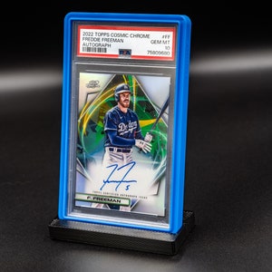 May include: A blue plastic graded card case with a baseball card inside. The card features a baseball player in a blue and white uniform holding a bat. The card is graded GEM MT 10 and has a signature on the bottom. The card is from the 2022 Topps Cosmic Chrome series and features Freddie Freeman.