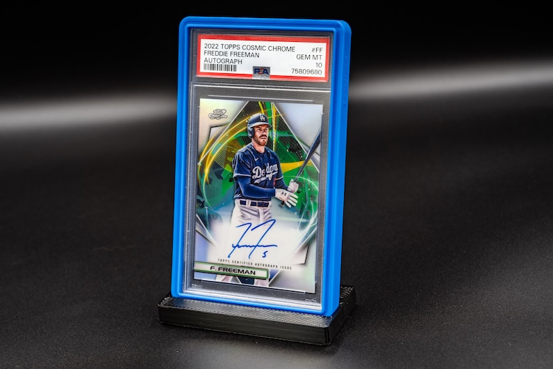 May include: A blue plastic graded card case with a baseball card inside. The card features a baseball player in a blue and white uniform holding a bat. The card is graded GEM MT 10 and has a signature on the bottom. The card is from the 2022 Topps Cosmic Chrome series and features Freddie Freeman.