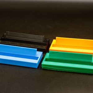 May include: Four plastic storage containers in different colors: black, blue, orange, and green. Each container has a rectangular shape with a raised lip on one side.