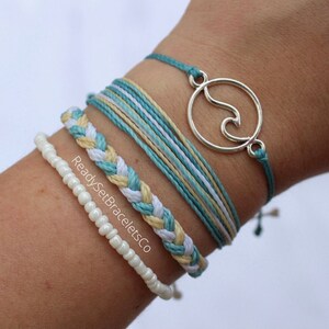 Puede incluir: Un conjunto de cuatro pulseras que incluyen una pulsera de cuentas blancas, una pulsera trenzada con hilos blancos y amarillos, una pulsera multi-hilo con hilos azules, blancos y amarillos, y una pulsera de cuerda azul con un dije de ola plateado. Las pulseras están apiladas en la muñeca de una persona.