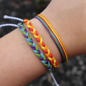 Puede incluir: Tres pulseras de amistad coloridas en la muñeca de una persona. Las pulseras están hechas de cuerda trenzada en colores arcoíris, incluyendo rojo, naranja, amarillo, verde, azul y morado.