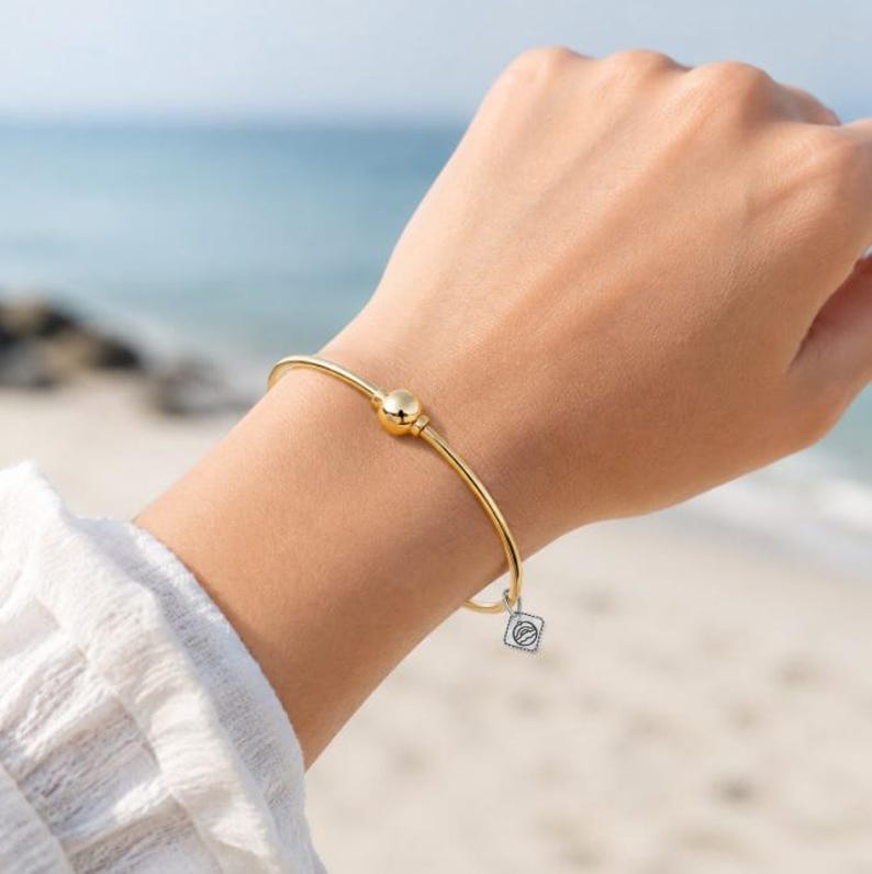 May include: A gold-toned bangle bracelet featuring a single centered spherical bead and a small, square-shaped silver-toned charm dangling from the band. The bracelet has a smooth, polished finish and a thin, circular wire construction.