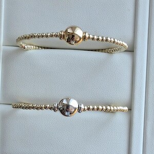 May include: Two gold-colored beaded bracelets. Each bracelet features a large, smooth, spherical bead at the center. The bracelets are displayed on a white surface. The beads are reflective, showing the surrounding environment.