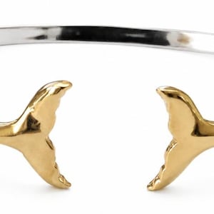 May include: A silver-toned cuff bracelet featuring two gold-toned whale tail charms at the open ends. The bracelet band is smooth and polished, with textured, rope-like silver detailing where the whale tails connect to the main band.