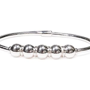 May include: A silver bangle bracelet with five round, polished beads. The bracelet has a simple, elegant design and a clasp closure. The beads are evenly spaced along the front of the bracelet.