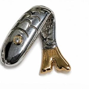 May include: A silver-toned metal brooch shaped like a curved fish with engraved scale patterns along its body. The fish features a single clear round gemstone set in a gold-toned circular bezel near the head and a polished gold-toned tail fin.