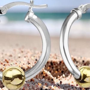 May include: A pair of silver hoop earrings with a gold-colored bead at the bottom. The earrings are displayed on a sandy surface with a blurred ocean background. The hoops have a polished finish.