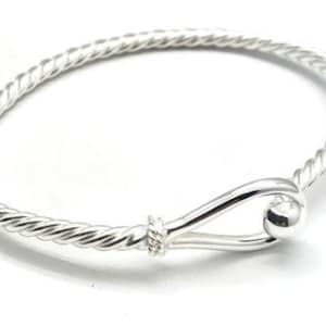 May include: A silver-colored bangle bracelet featuring a twisted rope-like texture along the band. The bracelet includes a hook-and-eye style closure mechanism with a smooth, polished spherical bead at the end of the hook, creating a continuous loop.