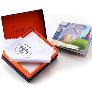 May include: A square jewelry box with a dark exterior and bright orange interior, containing a white polishing cloth with the black Cape Cod Jewelry logo. Beside the box is a small booklet featuring a coastal illustration and the same circular logo.