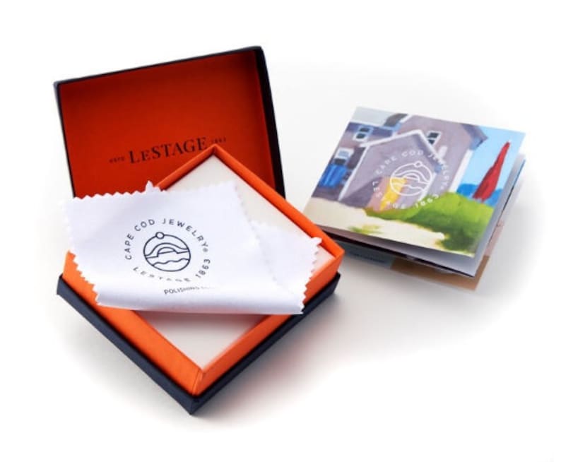 May include: A square jewelry box with a dark exterior and bright orange interior, containing a white polishing cloth with the black Cape Cod Jewelry logo. Beside the box is a small booklet featuring a coastal illustration and the same circular logo.