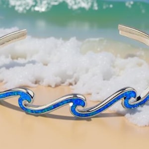 May include: Silver-toned cuff bracelet with a wave design. The bracelet features a series of blue, opal-like wave patterns. The open-ended bracelet is displayed on a sandy surface with a blurred ocean background.
