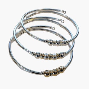 May include: A stack of silver-toned metal bangles with gold and silver beads. The bracelets have a simple, elegant design, with a curved shape and a series of small, round beads clustered in the center. The bangles are displayed against a white background.