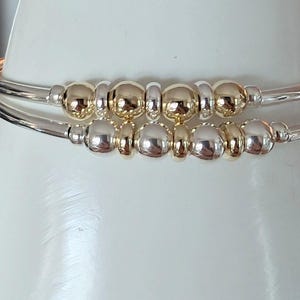 May include: Two silver bangles with gold and silver beads. The bracelets are stacked, with the top one featuring gold beads and the bottom one silver. The beads are evenly spaced along the center of the bracelets.
