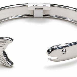 May include: A silver-toned metal cuff bracelet featuring a fish-shaped design. One end of the bracelet is shaped like a fish tail with etched lines, while the other end depicts a fish head with a small embedded clear stone for an eye.