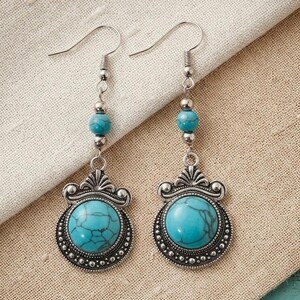 May include: A pair of dangle earrings featuring a round, turquoise-colored stone with dark veining set in an ornate, antiqued silver-tone metal frame. Each earring includes a smaller matching bead above the main pendant and a silver hook wire.