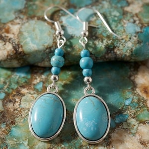 May include: A pair of dangle earrings featuring oval-shaped turquoise stones set in silver-toned metal frames. Each earring includes two small round turquoise beads and a single silver-toned spacer bead suspended from a silver-toned hook wire.
