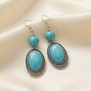 May include: A pair of drop earrings featuring oval-shaped turquoise-colored stones with dark veining, set in silver-toned metal frames with a textured border. Each earring includes a round turquoise-colored bead above the oval pendant and a hook.