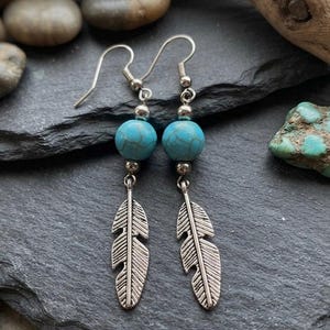 May include: A pair of dangle earrings featuring a single round turquoise-colored bead with dark veining, accented by small silver-toned metal beads and a silver-toned metal feather charm suspended from a standard silver-toned ear wire hook.