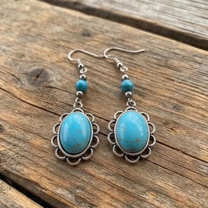 May include: A pair of dangle earrings featuring oval-shaped turquoise-colored stones set in scalloped silver-toned metal frames. Each earring includes a small round turquoise-colored bead above the main setting, suspended from a silver-toned hook wire.