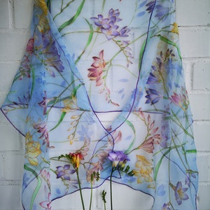 以下が含まれることがあります： 紫、ピンク、黄色の花柄が特徴の、透明感のある水色のスカーフ。紫色の縁取りが施された、繊細な水彩画のようなデザインです。白いレンガの壁を背景に、白いハンガーに掛けられています。