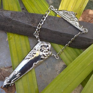 May include: A silver-toned, ornate, decorative scissor holder with a chain and a floral design. The holder is shaped like a knife and has a pointed tip.