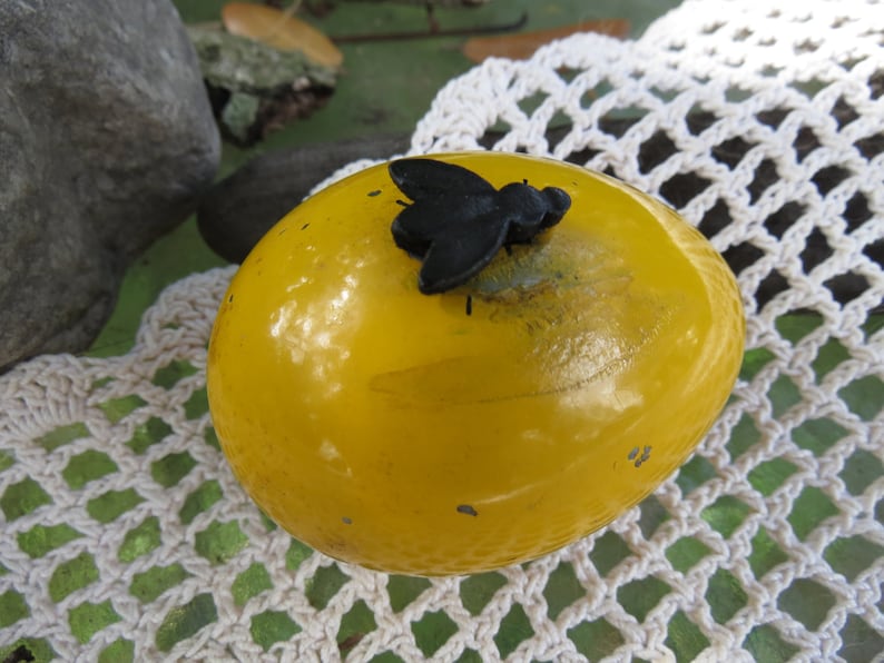 Pu&ograve; includere: Un uovo giallo con una mosca di plastica nera sopra. L'uovo &egrave; su una superficie di uncinetto bianca.
