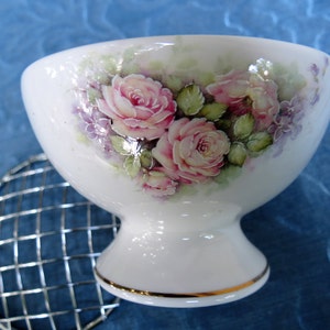 Puede incluir: Cuenco de cer&aacute;mica blanco con pie, con un dise&ntilde;o floral con rosas rosas y detalles morados. El borde est&aacute; adornado con oro. Una ranita de metal plateado est&aacute; al lado del cuenco.