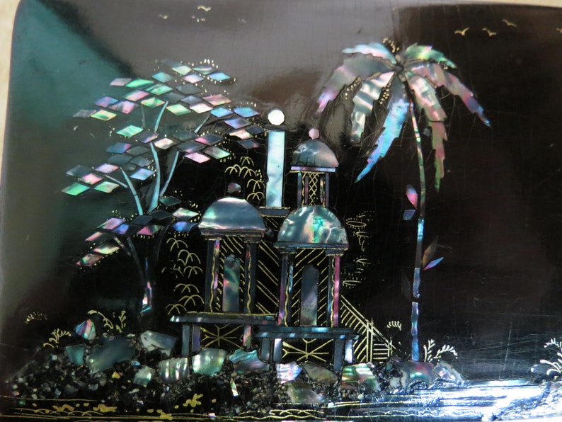 Pu&ograve; includere: Una scatola laccata nera con una scena di un pagoda e palme intarsiate con madreperla iridescente. La scena &egrave; delineata con vernice dorata.