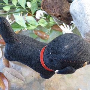 May include: A handmade black cat-shaped stuffed animal with a red collar. The cat is made of a dark fabric with visible stitching in a contrasting color. The tail is upright, and the cat is lying down.