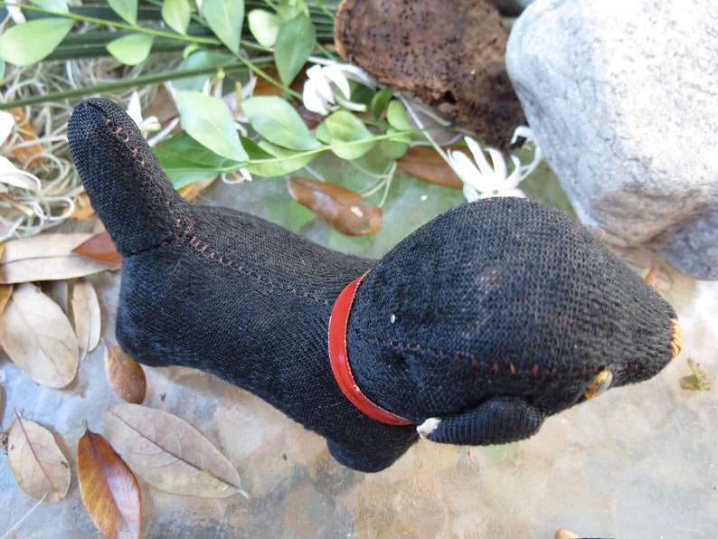 May include: A handmade black cat-shaped stuffed animal with a red collar. The cat is made of a dark fabric with visible stitching in a contrasting color. The tail is upright, and the cat is lying down.