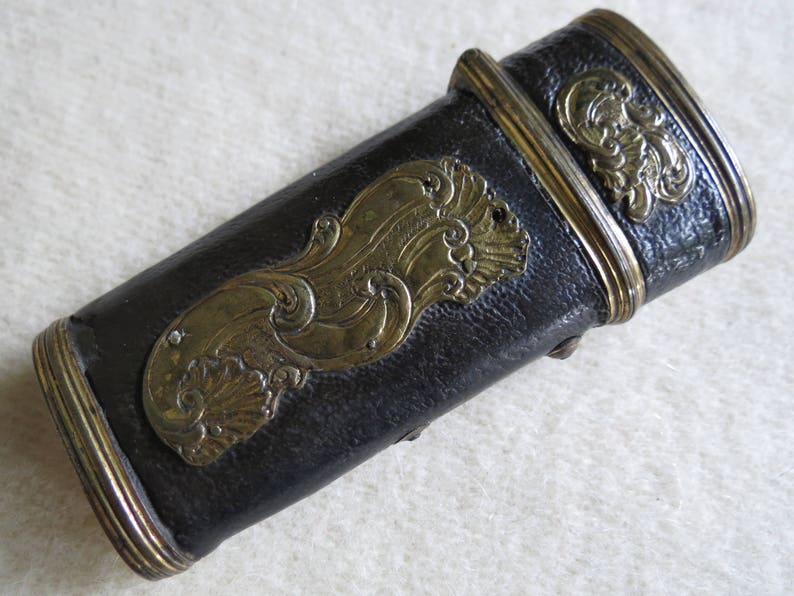 Op de afbeelding: Een zwart leren etui met gouden metalen accenten en een decoratief ontwerp. Het etui heeft een scharnierend deksel en een decoratieve metalen rand rond de randen.