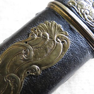 Op de afbeelding: Close-up van een zwart leren schede met een gouden metalen decoratieve rand. De rand heeft een golvend ontwerp met ingewikkelde details.