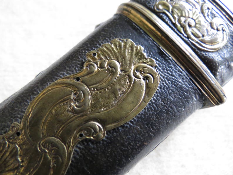 Op de afbeelding: Close-up van een zwart leren schede met een gouden metalen decoratieve rand. De rand heeft een golvend ontwerp met ingewikkelde details.