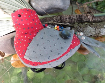 May include: A handmade fabric bird with a red head and body, patterned with white polka dots. The wings are gray with a small floral pattern. The bird has a large black button eye and is decorated with colorful beads and pearls.