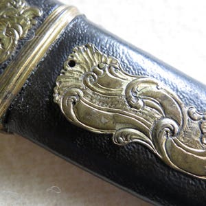 Op de afbeelding: Close-up van een zwart leren schede met gouden metalen accenten. De schede heeft een decoratief ontwerp met ingewikkelde bloemmotieven.