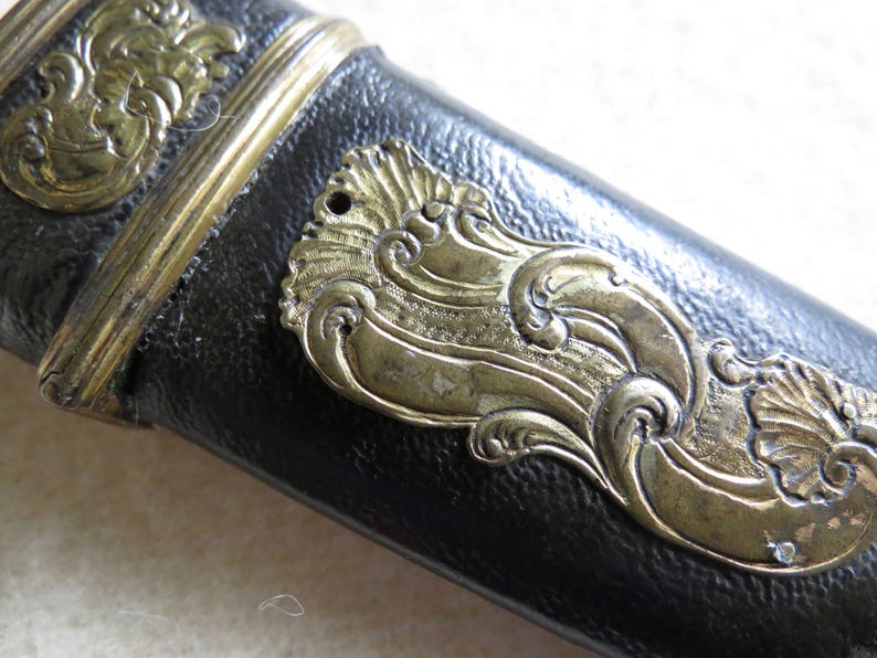 Op de afbeelding: Close-up van een zwart leren schede met gouden metalen accenten. De schede heeft een decoratief ontwerp met ingewikkelde bloemmotieven.