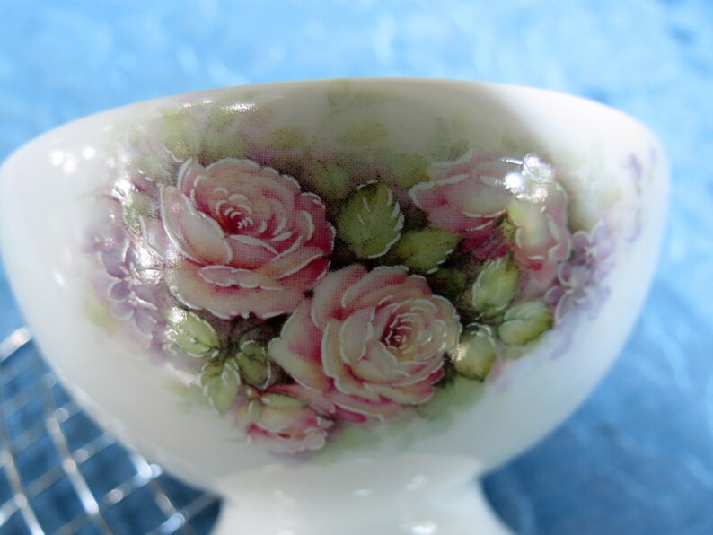 Puede incluir: Una taza de t&eacute; de porcelana blanca con base de pedestal. La taza est&aacute; decorada con un dise&ntilde;o floral pintado a mano con rosas rosas, flores moradas y hojas verdes. El fondo es un verde suave.