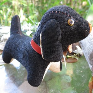 May include: A handmade black dog plush toy with a red collar. The dog has tan accents on its ears and paws, and a stitched orange nose. The eyes are large, round, and amber-colored. The plush is sitting on a reflective surface.