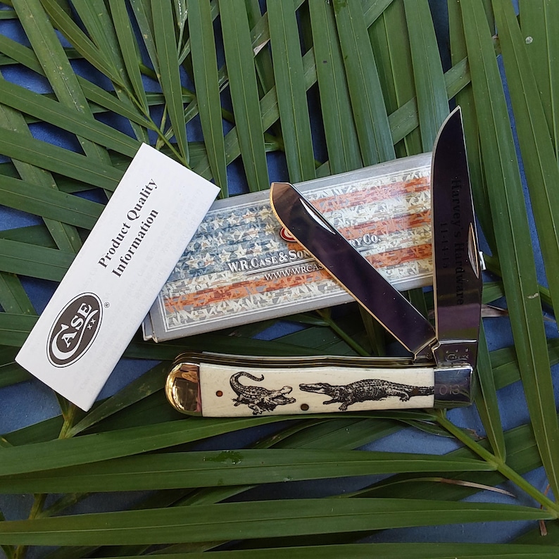 May include: A Case XX pocket knife with a bone handle featuring an alligator design. The knife is open and the blade is made of stainless steel. The knife is packaged in a box with a red, white and blue American flag design.