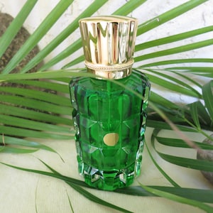 May include: A green glass perfume bottle with a gold-colored cap. The bottle has a geometric design and a small gold label. The background includes green leaves and a white wall.